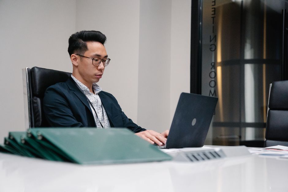 Asian man in suit working on a laptop at an office desk, surrounded by documents.